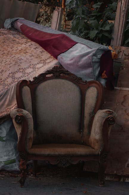 A broken and stained upholstered sofa resting outdoors on a sidewalk next to a paved pathway. The sofa has a tufted, dusty pink fabric that is torn and soiled, with stuffing and fabric fibers exposed 