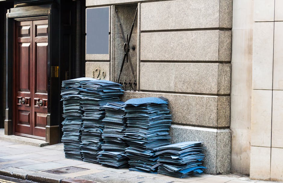 Stacks of disorganized blue file folders and documents piled against a stone and brick wall outside a building, near a wooden door and a large outdoor clock. The folders appear to be weathered, with s